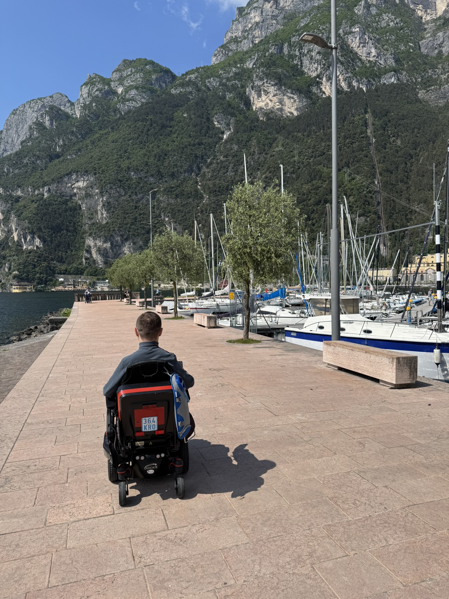 Andrei fährt mit seinem Elektrorollstuhl eine Hafenpromenade entlang. Vor ihm liegen der See, Segelboote und Berge.
